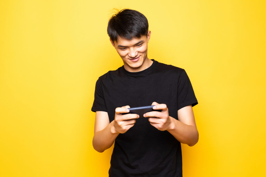 Young asian man play games by mobile phone standing isolated over yellow background