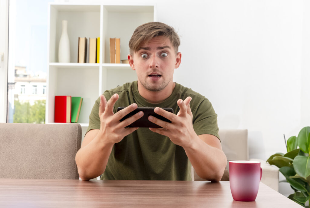 surprised-young-blonde-handsome-man-sits-table-with-cup-holding-looking-phone-inside-living-room